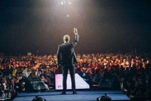 A back view of a man on stage waving to a large engaged crowd at a seminar. Symbolising the transition from a life of proving to a life of pride and purpose.